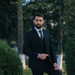 A sophisticated man in a suit posing confidently in a lush outdoor garden.